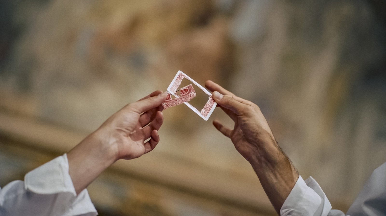 Le mani di due illusionisti si passano una carta attraversata da un&rsquo;altra carta, davanti a una parete affrescata